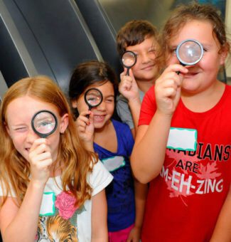 A group of children peer through magnifying glasses at a community activity.